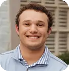 A person with short curly hair smiles at the camera. They are wearing a light blue, collared shirt and a zip-up jacket. The background is blurred with a structure featuring horizontal lines.