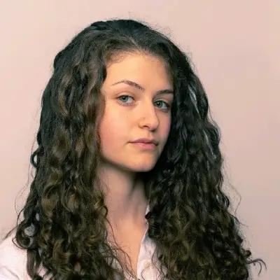 Person with long, curly hair and a neutral expression, wearing a light shirt, against a beige background.