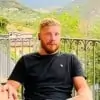 A person with short blonde hair and a beard sits in an outdoor setting, wearing a black t-shirt. In the background, there is a railing and green foliage, with hills and a partly cloudy sky visible.